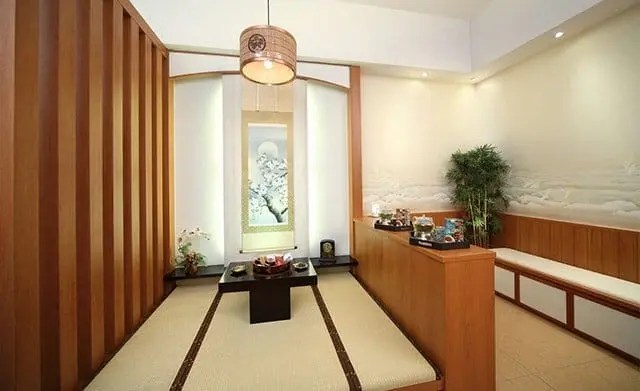 A minimalist Japanese-style tea room with tatami mats, a low table, wooden paneling, bench seating, and a decorative scroll on the wall.