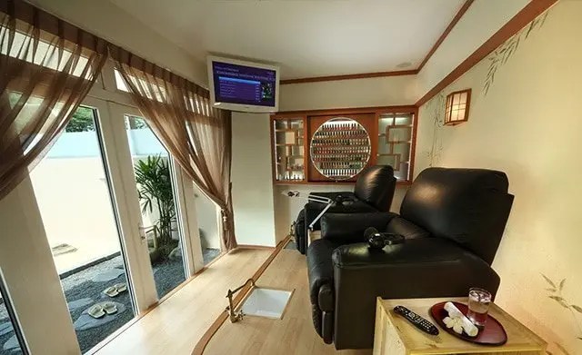A cozy spa room with two black recliner chairs, a TV on the wall, shelves with nail polish bottles, and large windows with sheer curtains.
