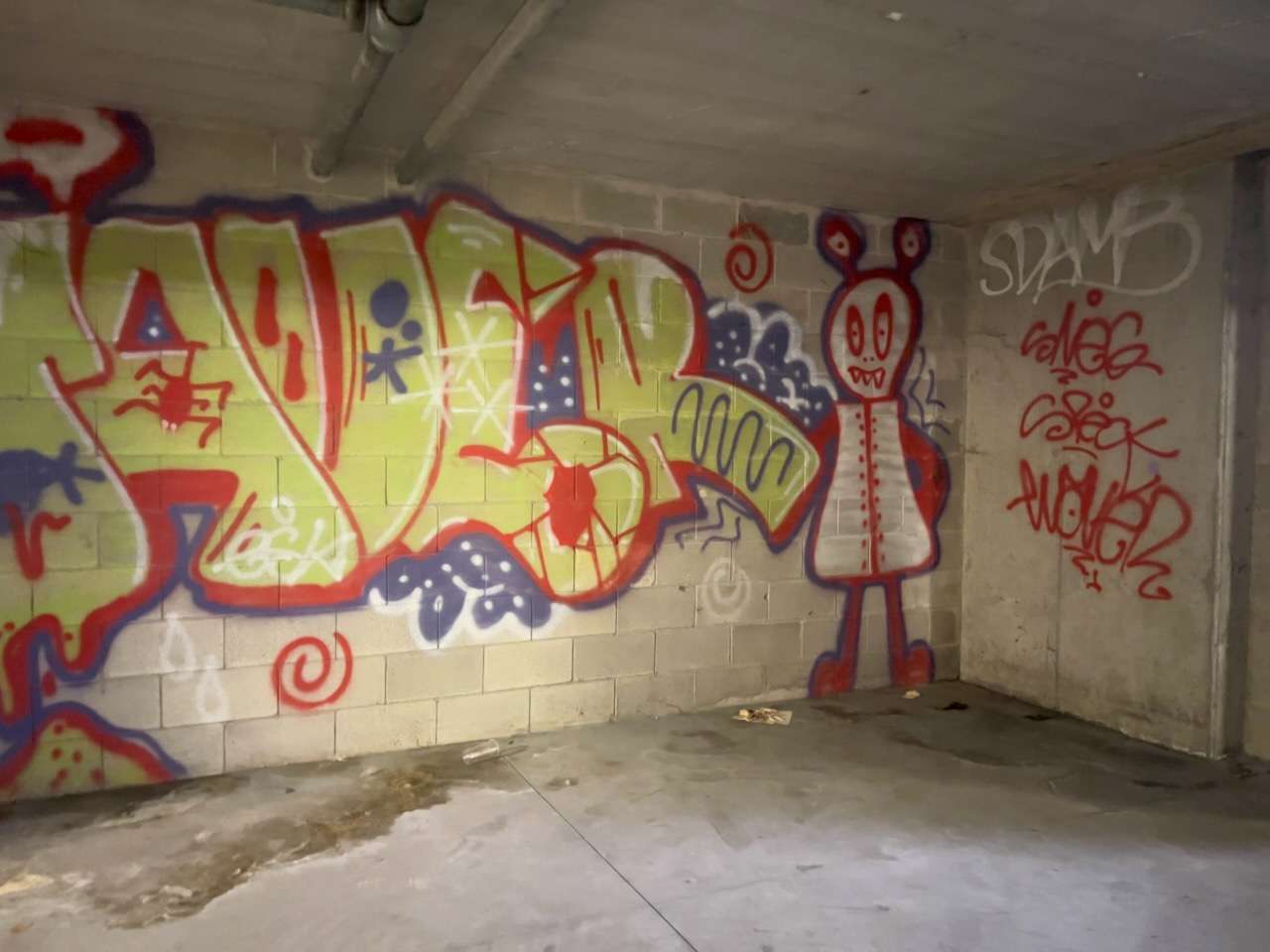 Graffiti colorati su un muro di blocchi di cemento in un locale abbandonato: grande scritta verde acido con contorni rossi e blu, un personaggio stilizzato rosso, alcune tag nell'angolo.