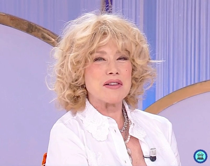 Woman with short curly blonde hair wearing a white blouse with a lace collar, speaking on a TV studio set with a lavalier mic clipped to her shirt.