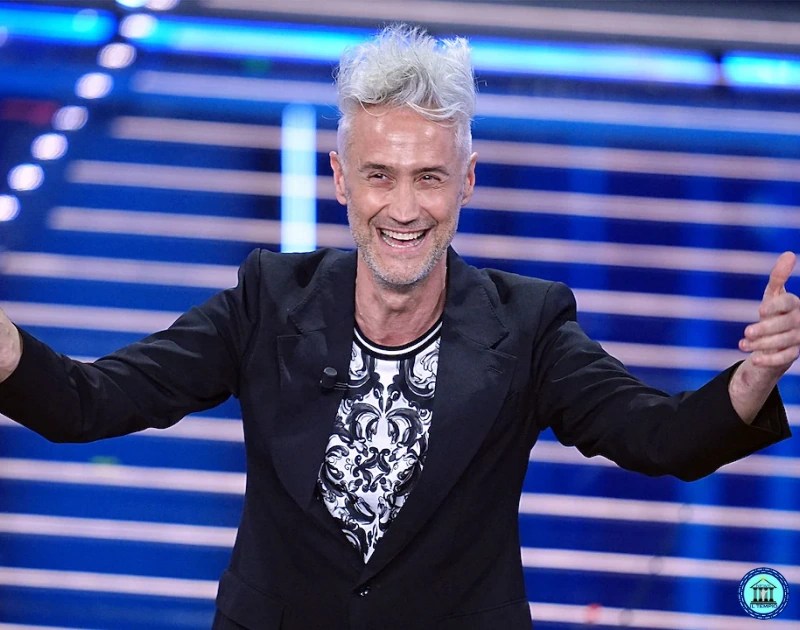 Smiling man with silver hair in a black blazer extends his arms on a blue-lit studio stage.