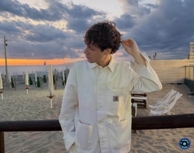 Young man in a white jacket on a sandy beach at sunset, looking to his left with one hand touching his hair.