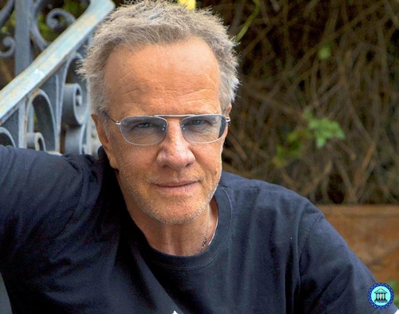 Casual outdoor portrait of a man with gray hair and sunglasses, wearing a black shirt, looking at the camera near a decorative railing and greenery in the background.