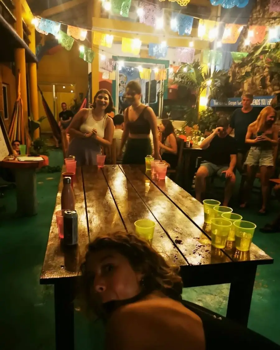 A girl leaning down under a camera while a photo is taken of people playing beer pong in a large courtyard with Mexican flags ahead and people enjoying themselves