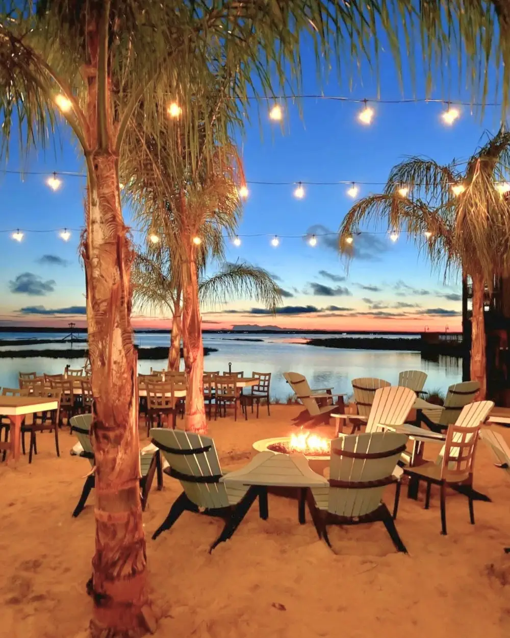 Adirondack chairs around a sand firepit under palm trees and string lights at sunset on the bay at Saltwater 75
