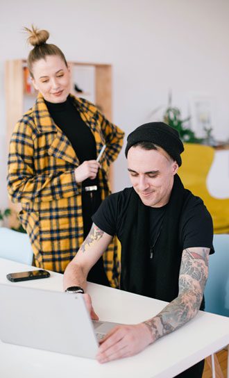 A person with tattoos and a black beanie is sitting at a desk using a laptop, perhaps working for a digital marketing agency in Hisar. Another person, wearing a yellow plaid coat and holding glasses, stands beside them, smiling. A mobile phone is on the table. The setting appears casual and creative.