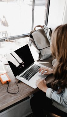 A person with long hair is sitting at a wooden table using a laptop, possibly working for a digital marketing agency in Hisar. A cup and an external hard drive are nearby. A backpack rests on the window ledge, and natural light pours through the window.