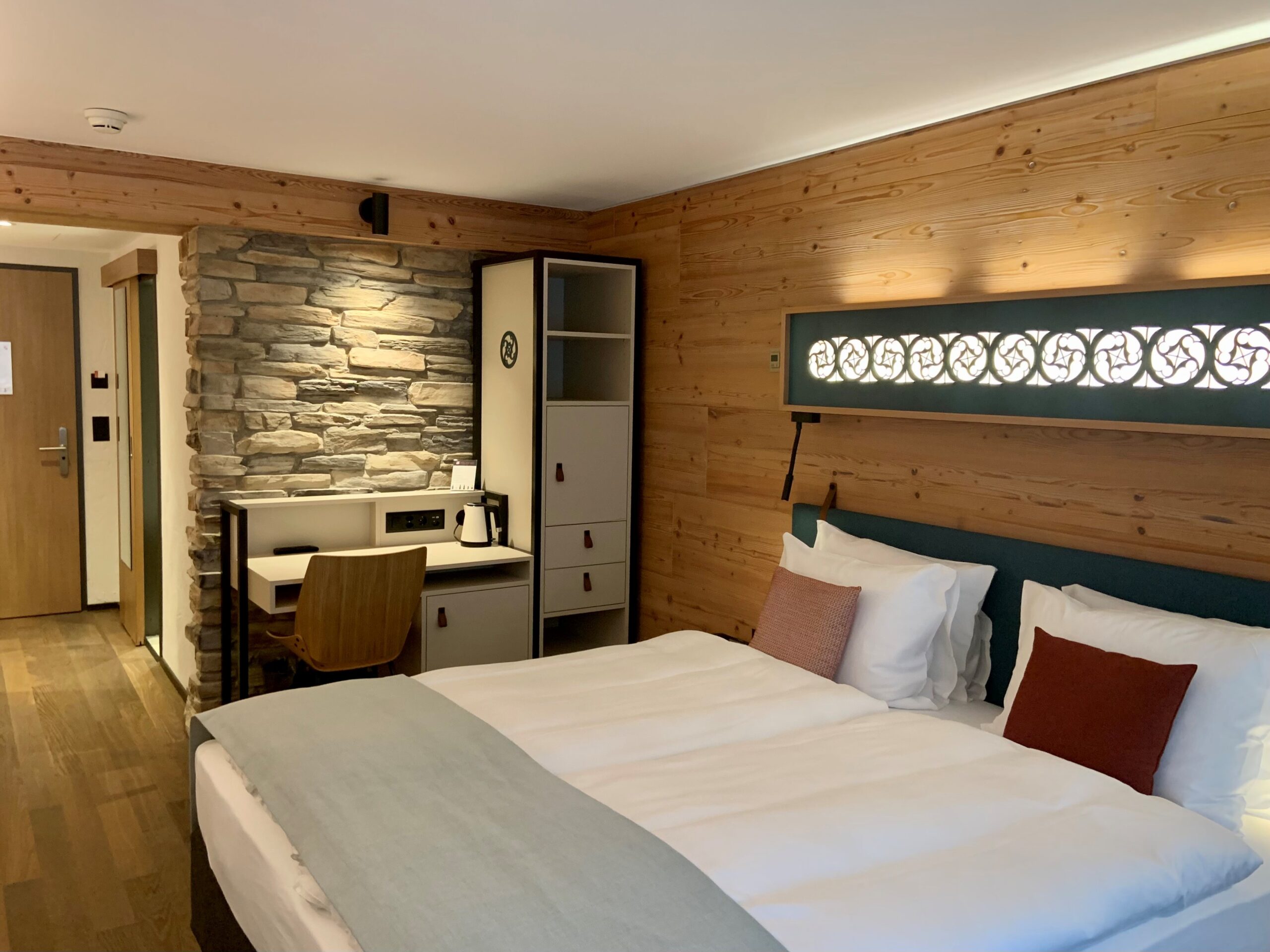 Ein gemütliches Hotelzimmer mit Doppelbett, weißer Bettwäsche und braunen Akzentkissen. Ein Holzschreibtisch mit Stuhl, ein Schrank und eine Steinwand verleihen dem Raum Charme. Warme Beleuchtung schafft eine einladende Atmosphäre.