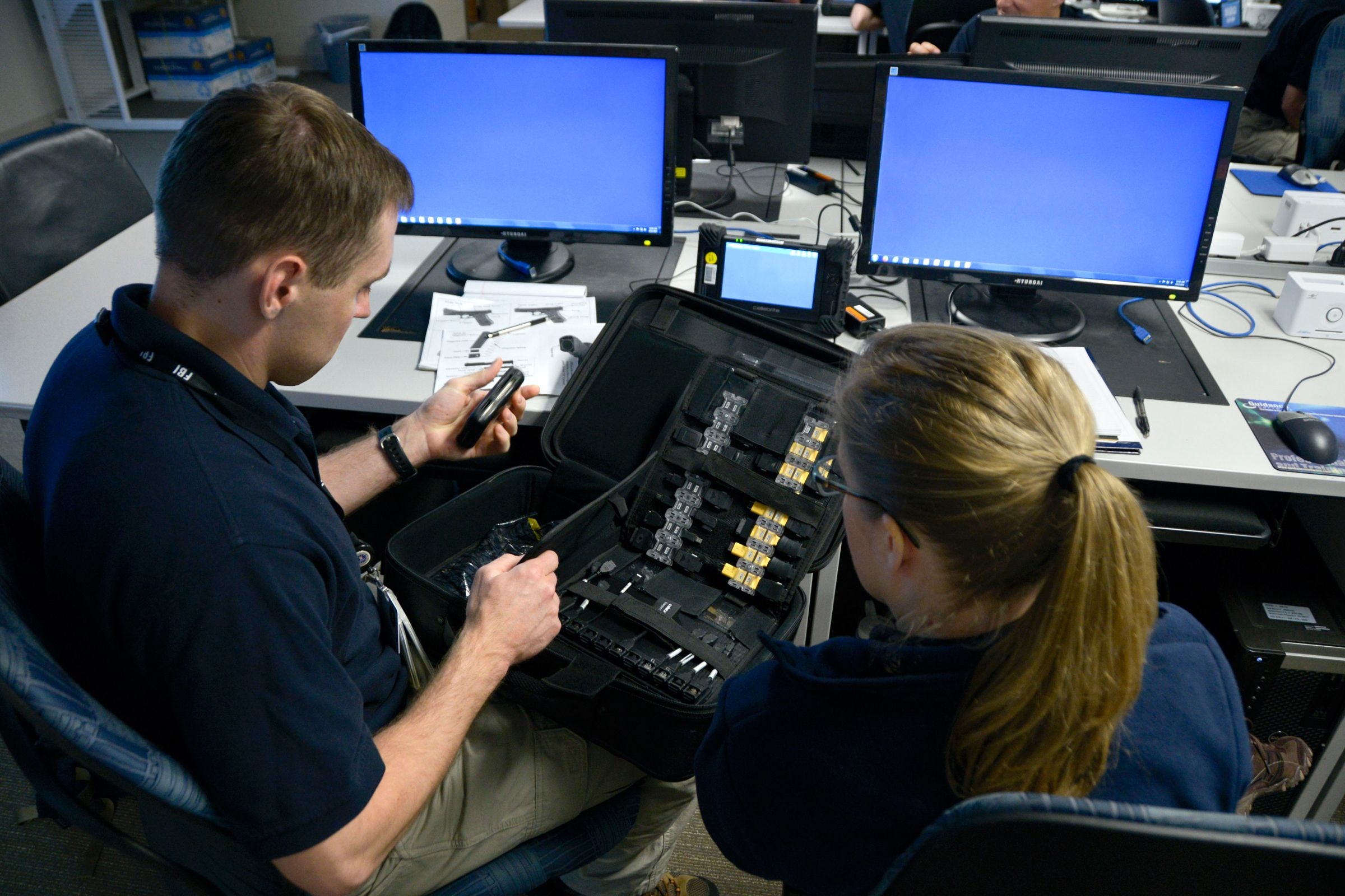 A FBI Agent and FBI Analyst sit a desk with computers and look at an open case with tools during a basic field training course.