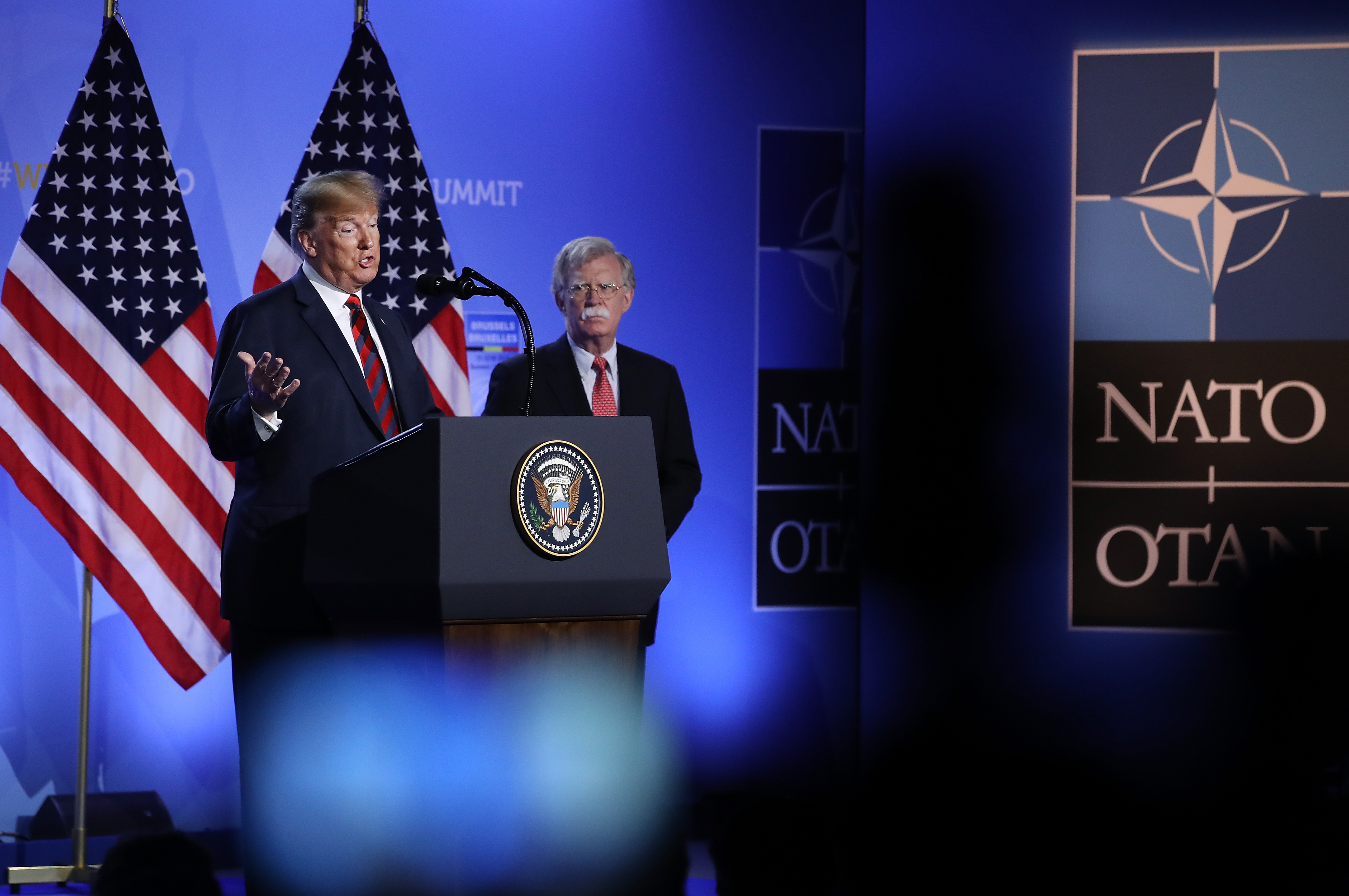 Trump, flanked by National Security Advisor John Bolton, speaks to the media at a press conference on the second day of the 2018 NATO Summit on July 12, 2018 in Brussels, Belgium. Two American flags stand behind them and behind that stands a backdrop with the NATO logo.