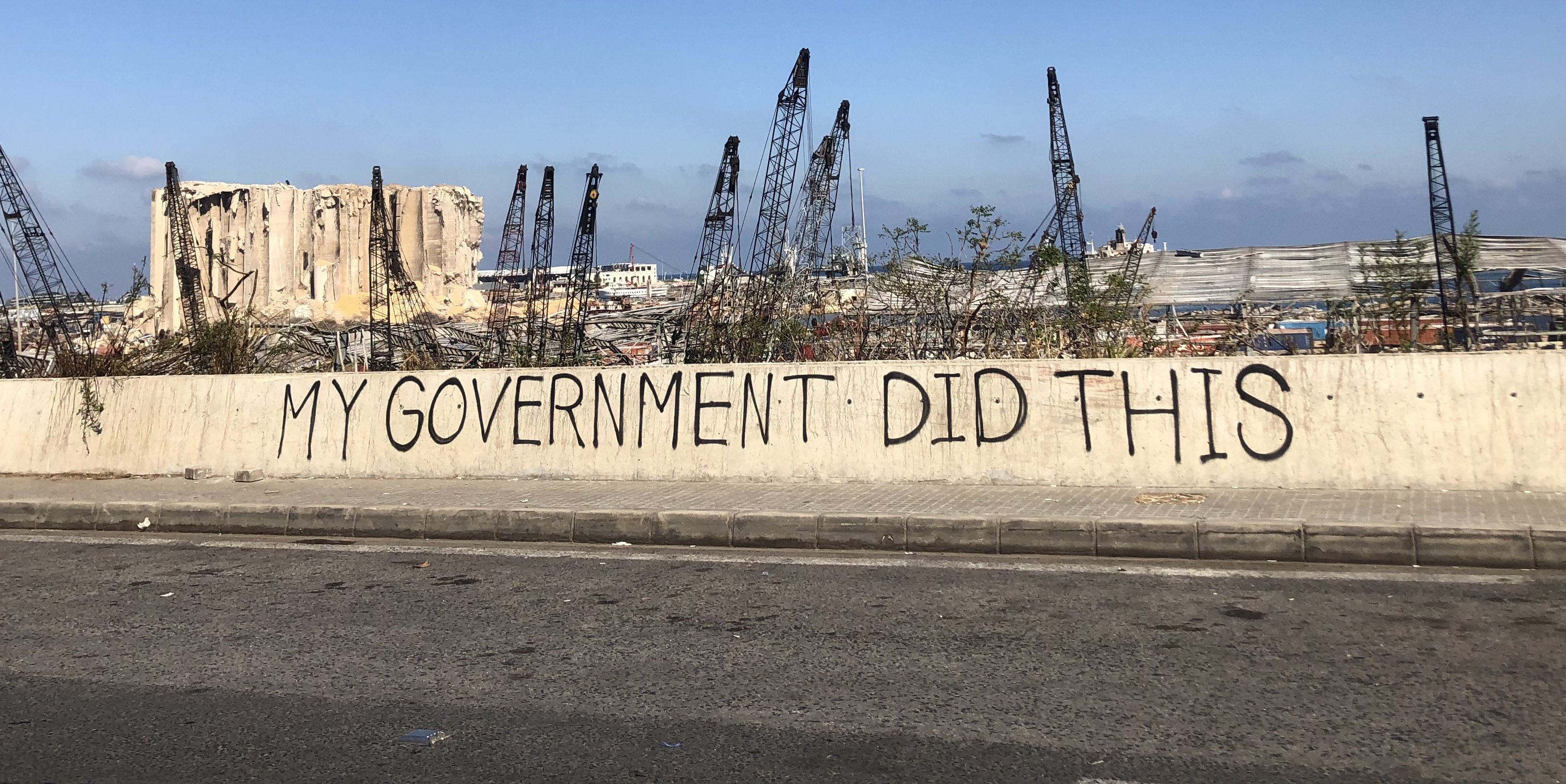 The words "My Government Did This" are displayed on a barrier on the Charles Helou highway in front of the ruined port of Beirut.