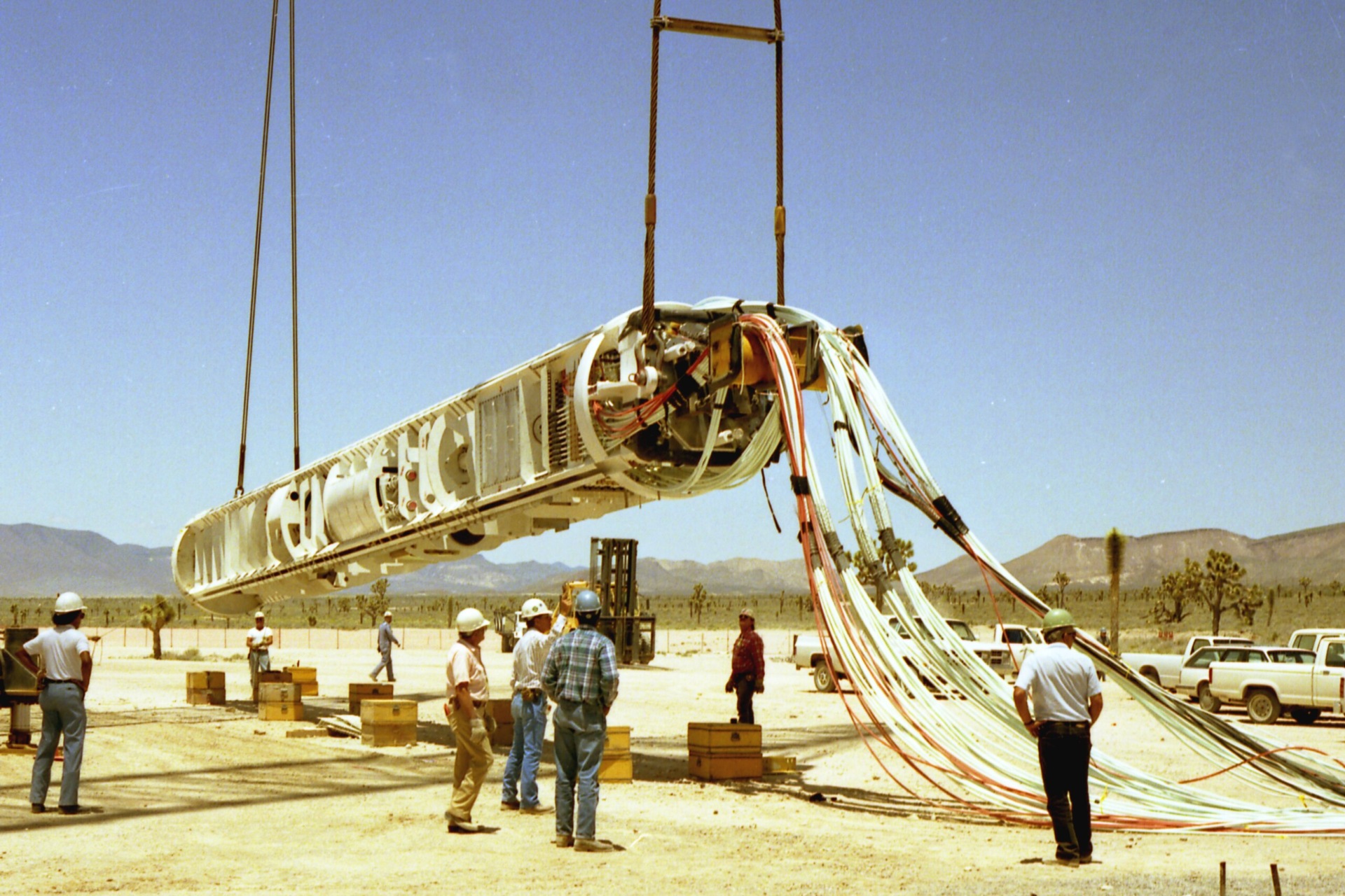 Workers wearing hard hats stand in a desert landscape under and around a long tube-like structure suspended from cables overhead. The tube appears to have differently sized and shaped compartments and equipment inside, and extending from the near end in the direction of the right side of the photo are numerous sets of cables in different colors, possibly connected to something offscreen.