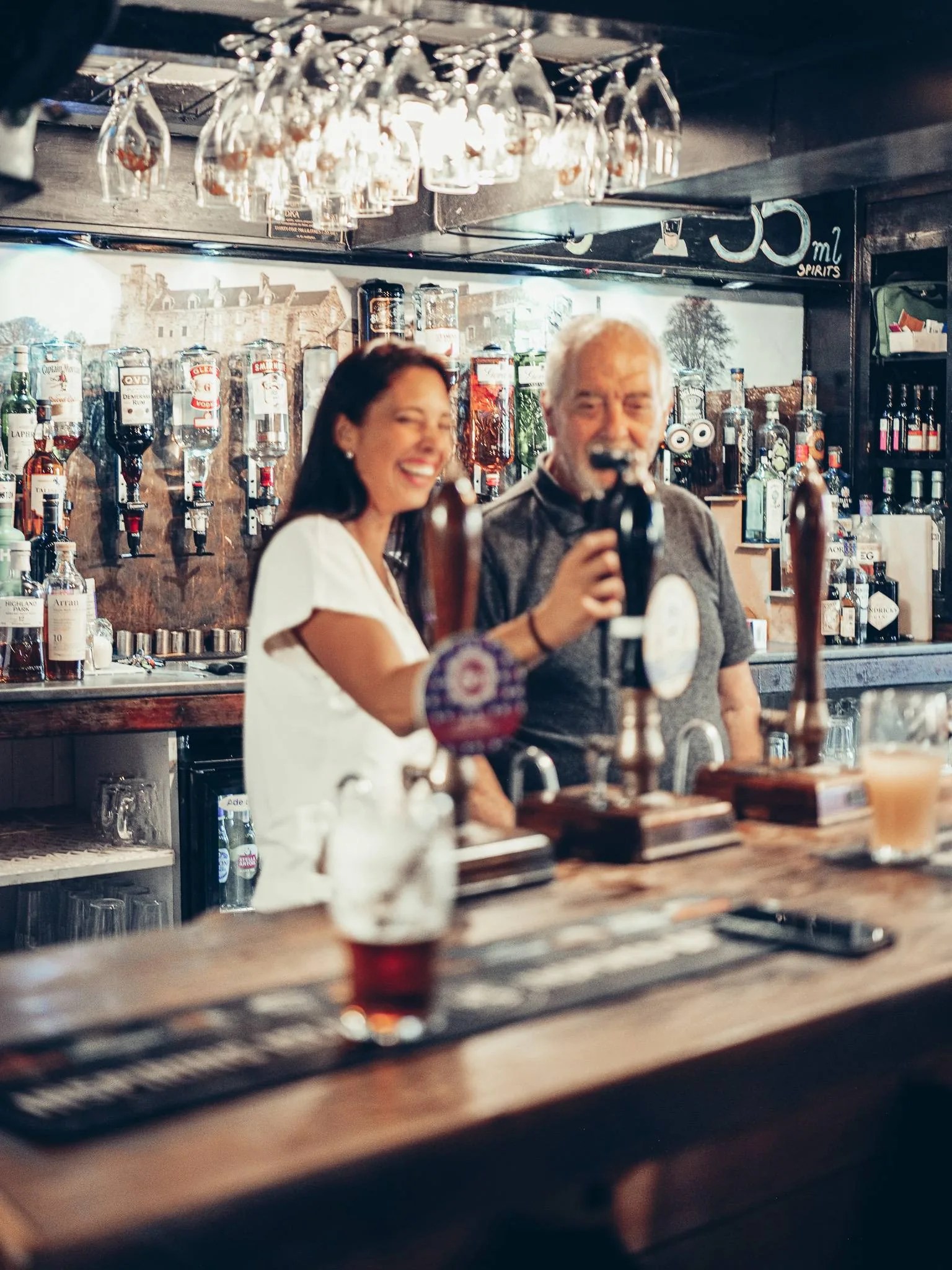 Caroline et Xavier au comptoir du Settle Inn, a Stirling, entoures des bouteilles et des verres suspendus au-dessus du bar.