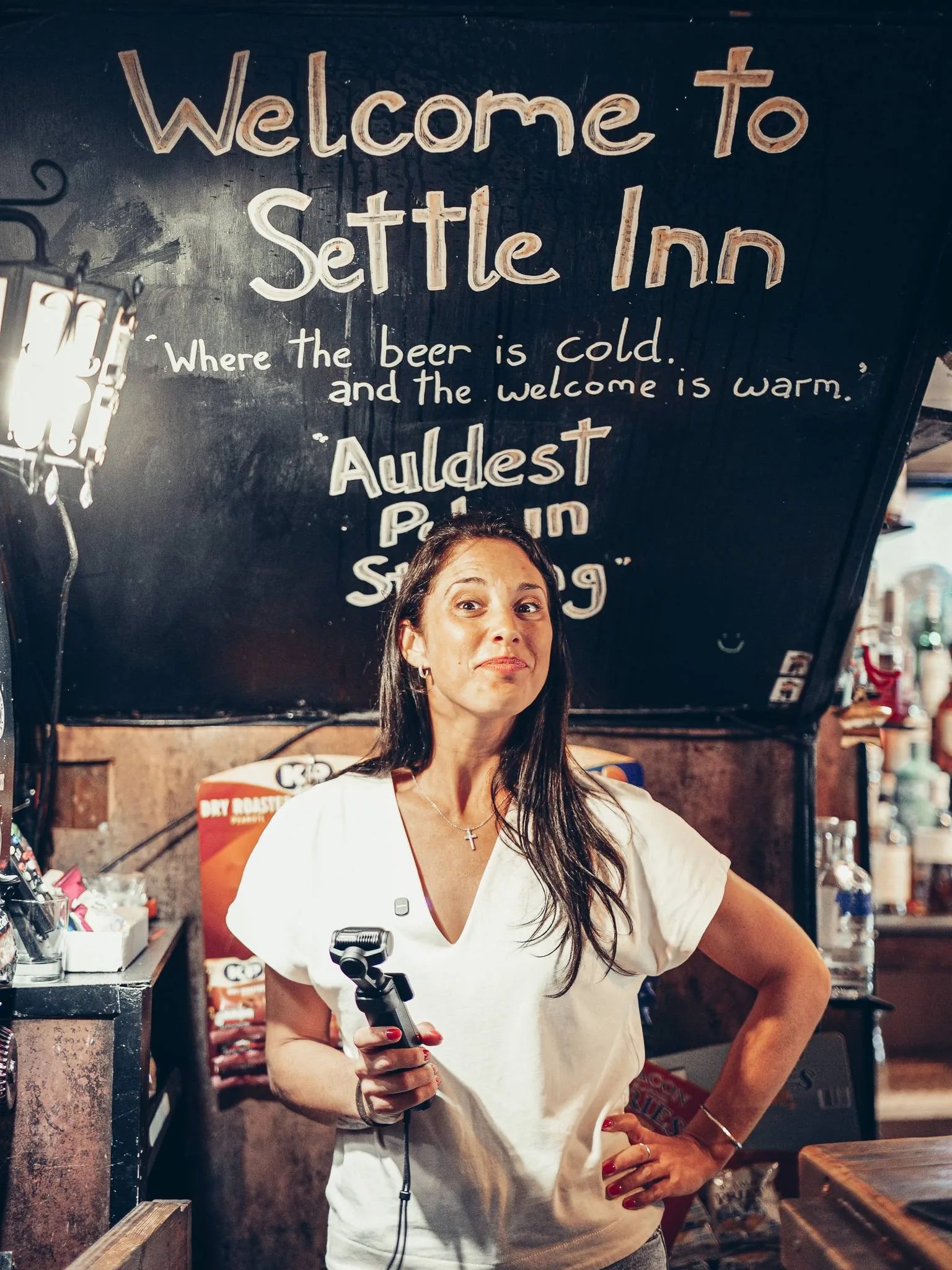 Caroline a l'interieur du Settle Inn, a Stirling, devant un panneau d'accueil, dans la penombre du pub.