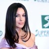 Monica Bellucci con capelli lisci e abito lilla scollato, indossa collana con ciondolo, posando con eleganza su sfondo con loghi Superfund.