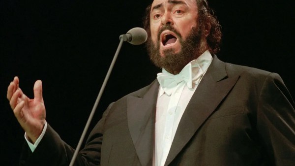 Luciano Pavarotti in giacca da sera nera e papillon bianco, canta appassionato di fronte a un microfono sul palco.