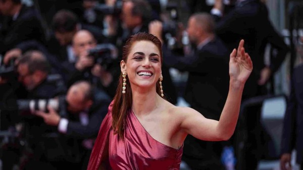 Donna in abito rosso monospalla che sorride e saluta con la mano alzata sul red carpet, fotografi in background