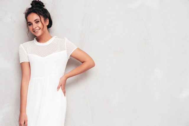 Giovane donna sorridente con capelli raccolti in uno chignon disordinato, indossa un abito bianco leggero con dettagli trasparenti a pois.