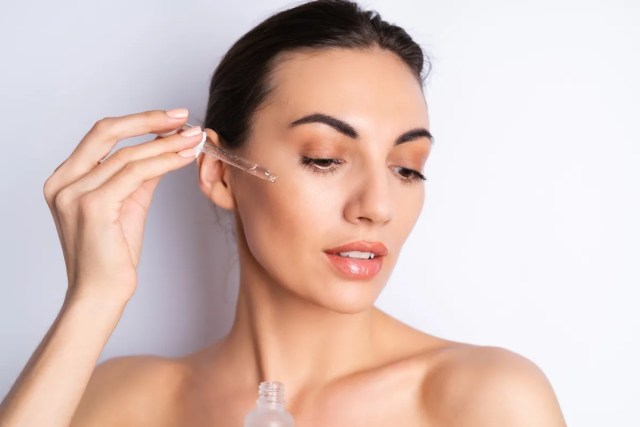 Giovane donna con pelle luminosa che utilizza un contagocce per applicare siero trasparente di acido ialuronico sul viso, come parte della sua routine skincare.