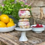Two ice cream sandwiches stacked on top of a small white cake plate sitting on a wooden table. Also pictured are a white bowl of lemons and a white bowl of raspberries.