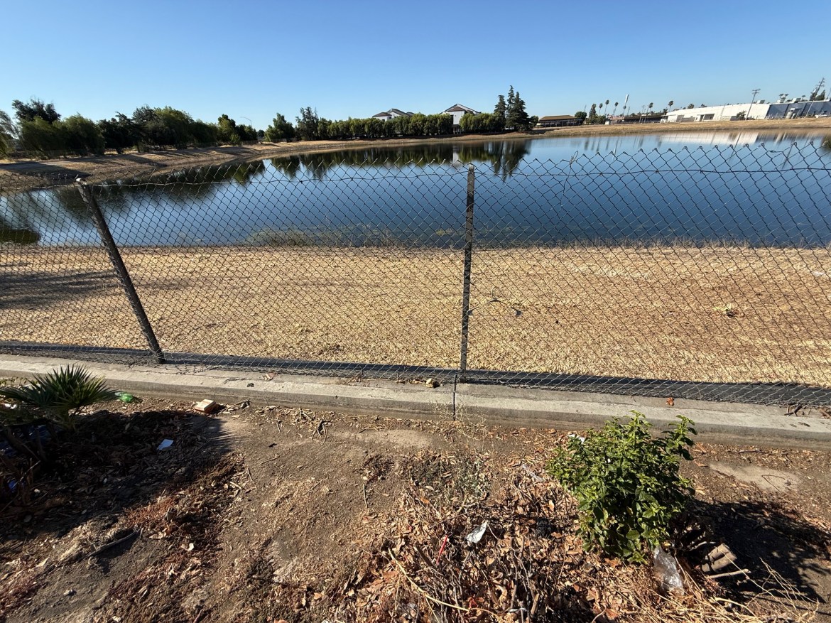 The same scene as the first image, but zoomed right out to show a background with an industrial unit of some kind next to the trees on the far bank, a tattered chain-link fence in front of the water, and waste ground in the foreground.