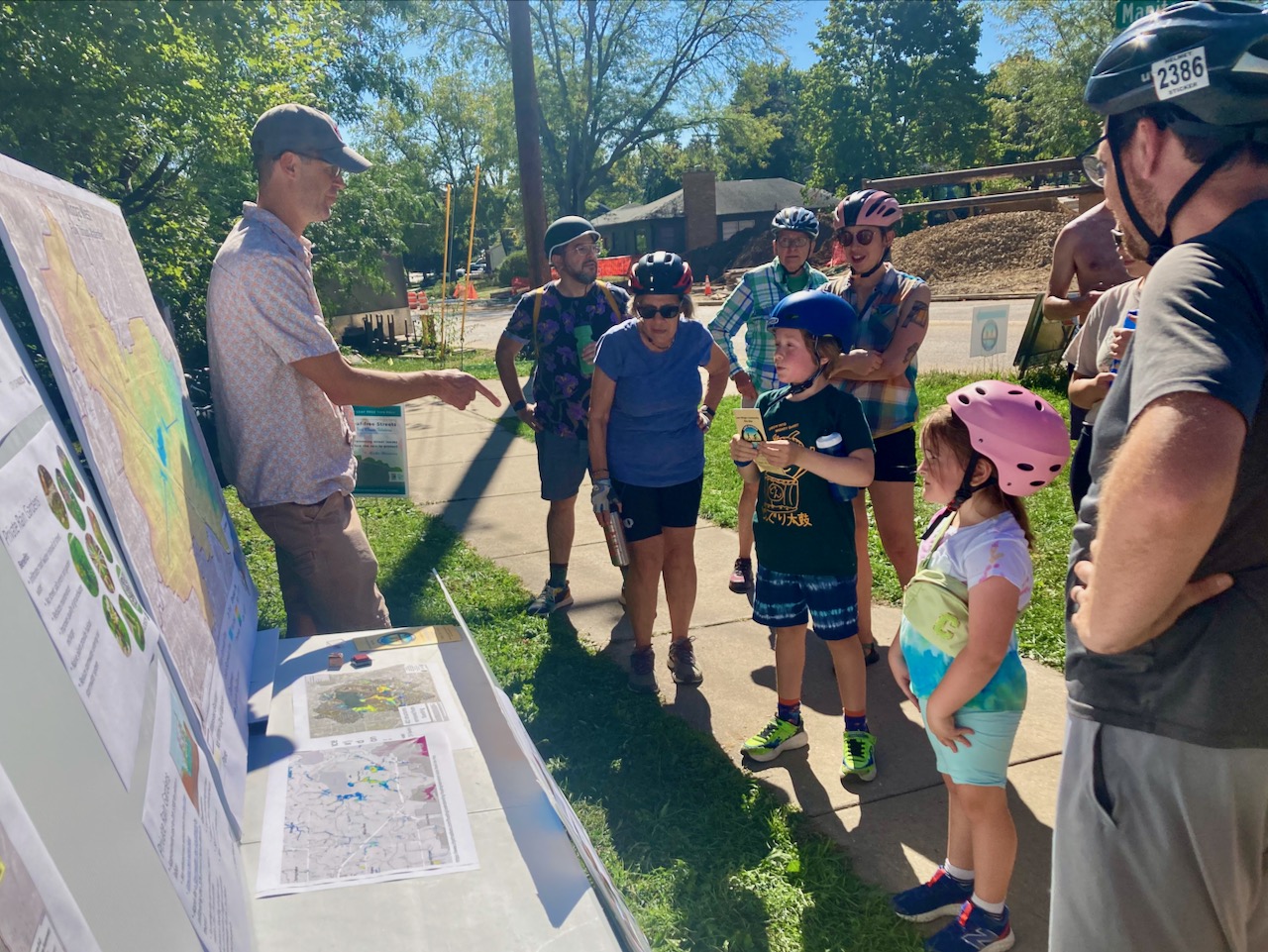 A group of people is facing a display and a person giving a presentation during the Lake Wingra Community Ride.