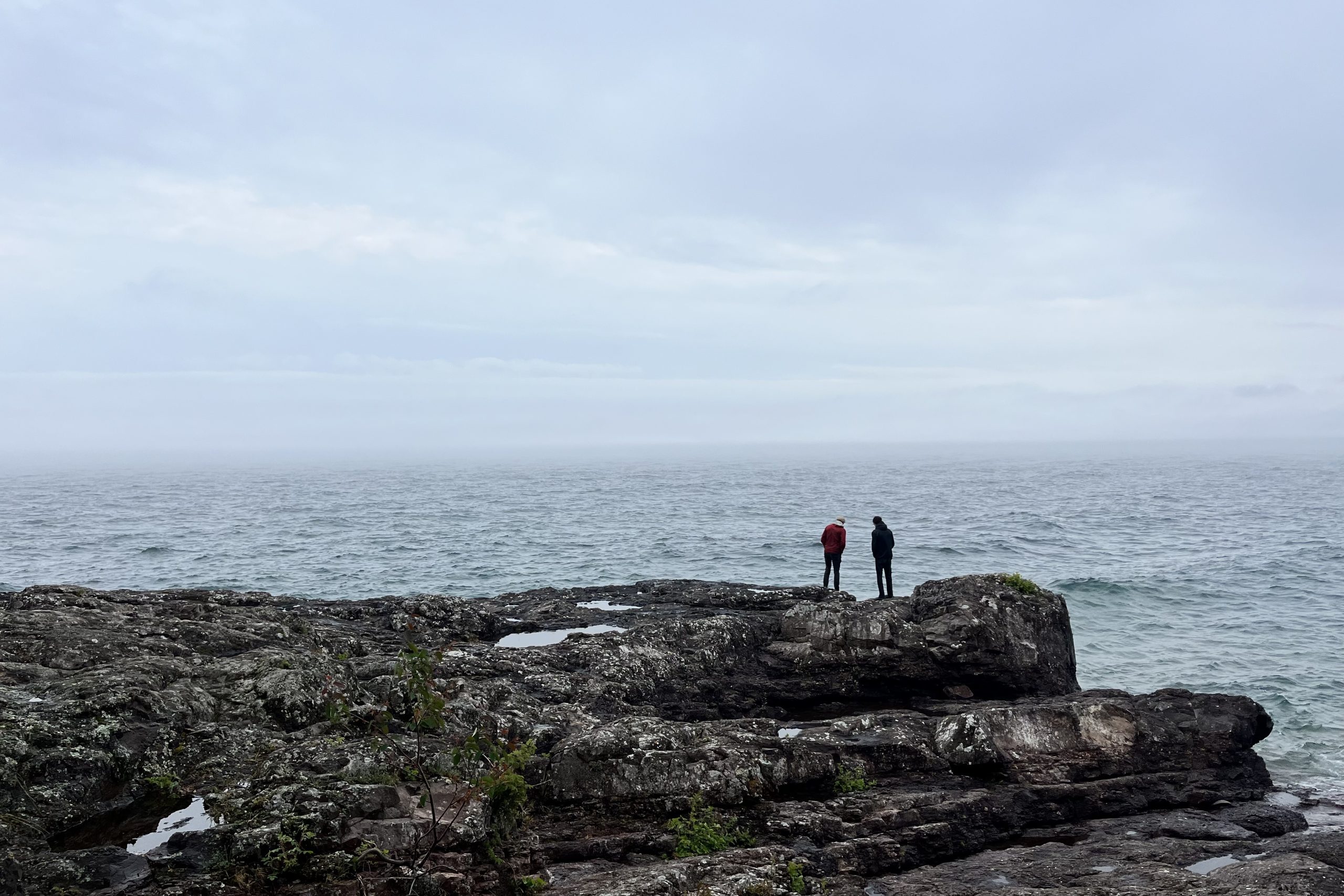 Two people in the distance standing on a slab of rock at the edge of a wide lake.