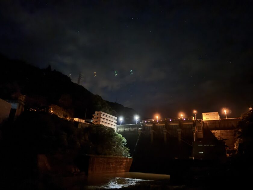 夜間にライトアップされた近代的なダムの堤体と、その周囲にある建物、上空に広がる星空