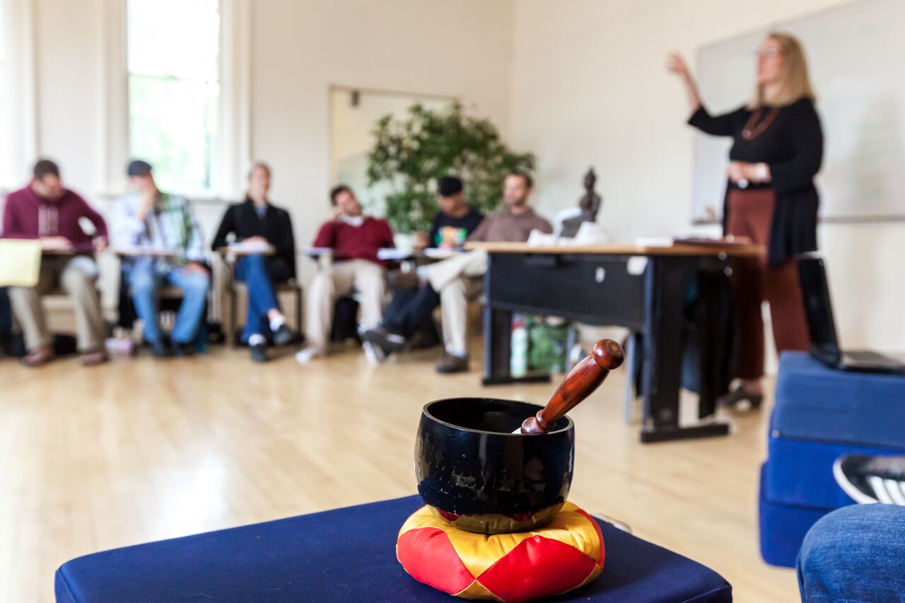 Meditation bowl in the foreground with a classroom of students and Judith Simmer Brown teaching in the background.