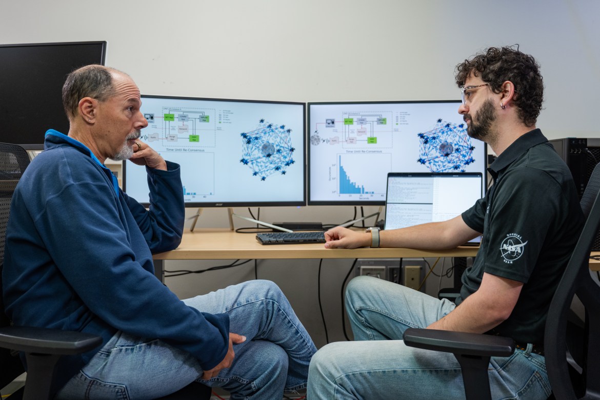 Two men sit conversing in front of a dual-screen monitor and laptop. The screens display spacecraft forming a network around the Moon.