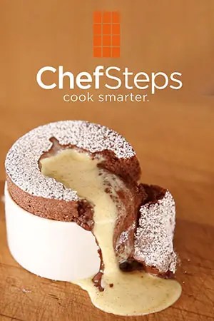 A chocolate soufflé topped with powdered sugar, partially cut open with vanilla sauce oozing out, sits on a wooden surface below the ChefSteps logo and tagline "cook smarter.
