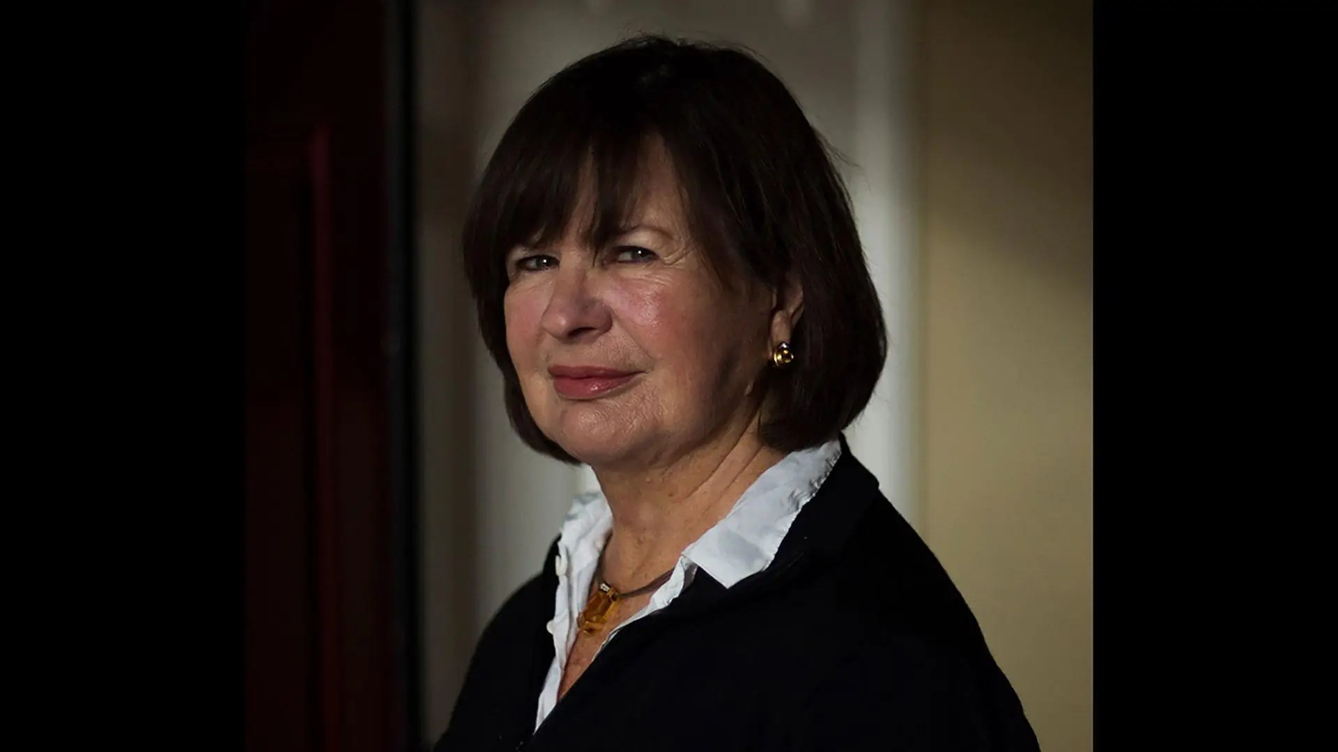 A woman with straight brown hair and a white-collared black top stands indoors, looking at the camera with a neutral expression.