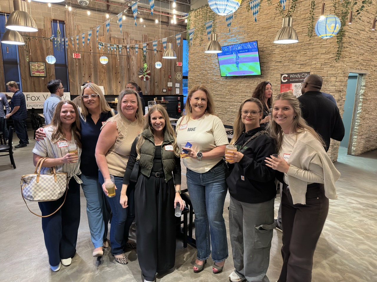 A group of seven attendees pose together with drinks, Fox Valley and Windy City Business Network banners behind them