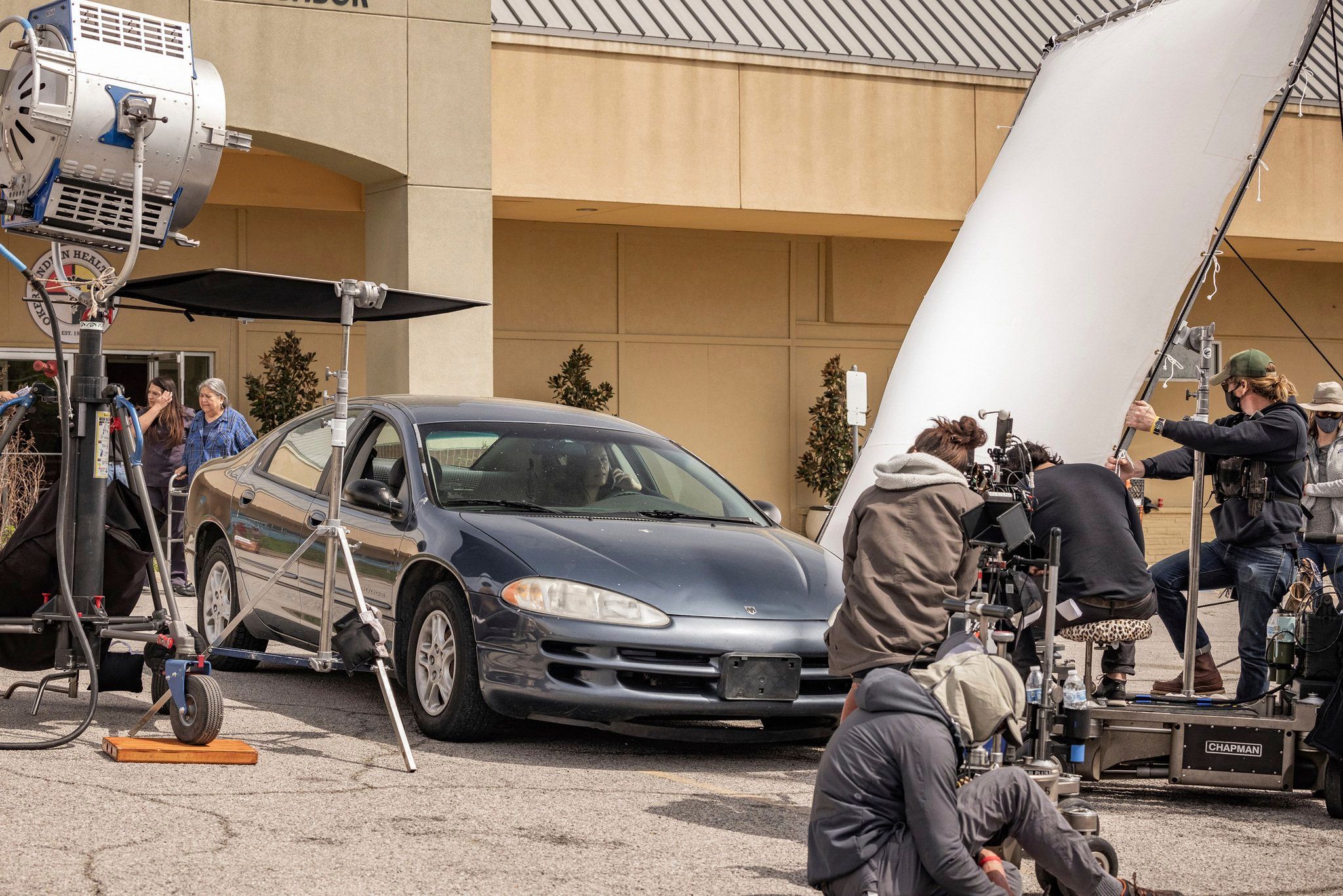 Behind-the-scenes view of a film set with film crew, lighting and camera equipment, a car and an actor inside the car.