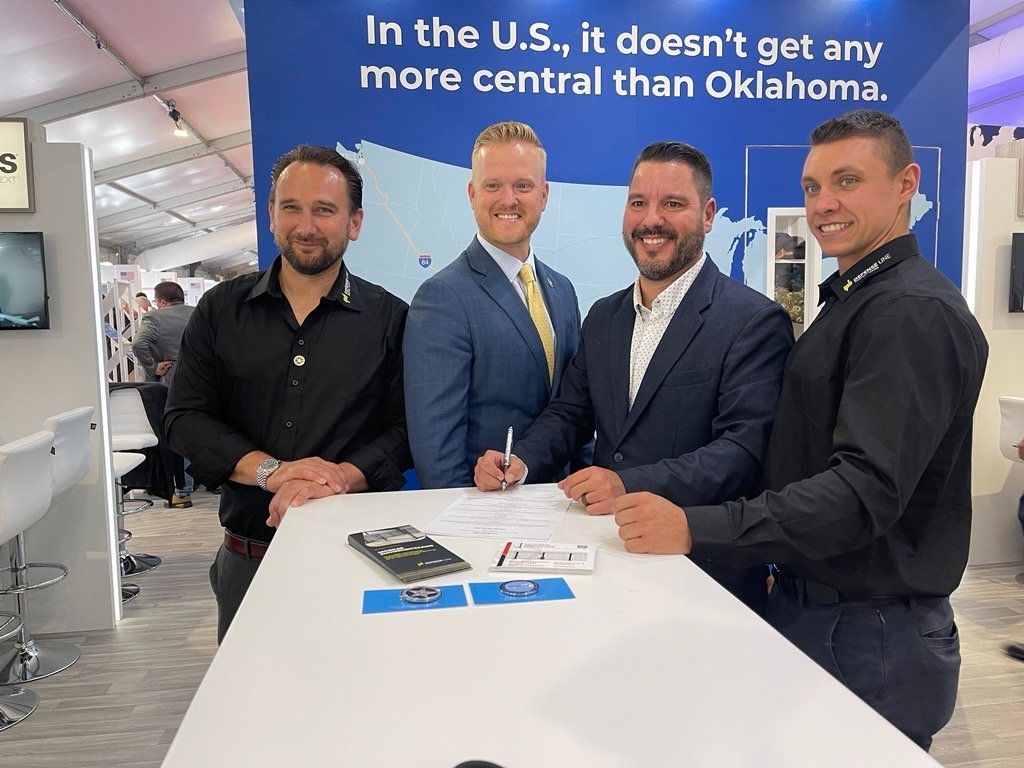 Four men standing in a row at a high table at a tradeshow with a banner in the background. One man holds a pen to sign a document.