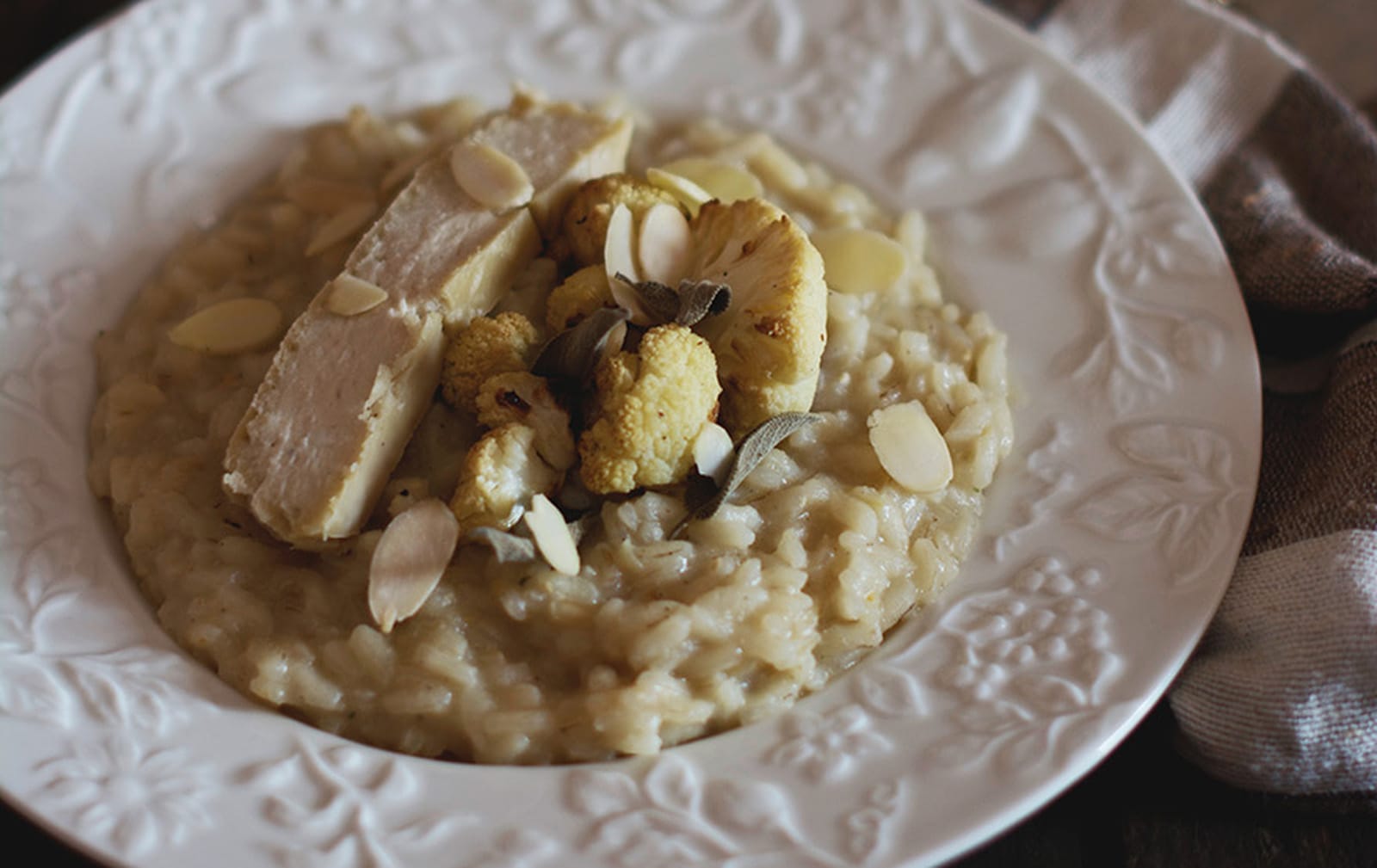 A bowl of creamy vegan cauliflower risotto, garnished with roasted cauliflower, fresh sage, a slice of fermented cashew cheese, and toasted almonds.