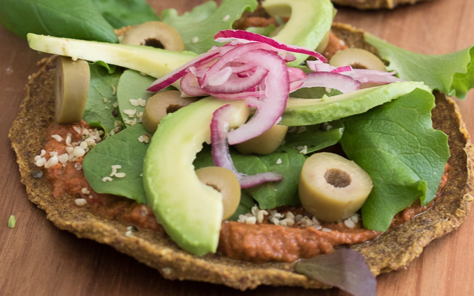 Plated raw sweet potato flatbreads topped with vibrant red tomato sauce, tangy pickled red onions, green olives, fresh arugula, and sliced avocado, ready to serve.