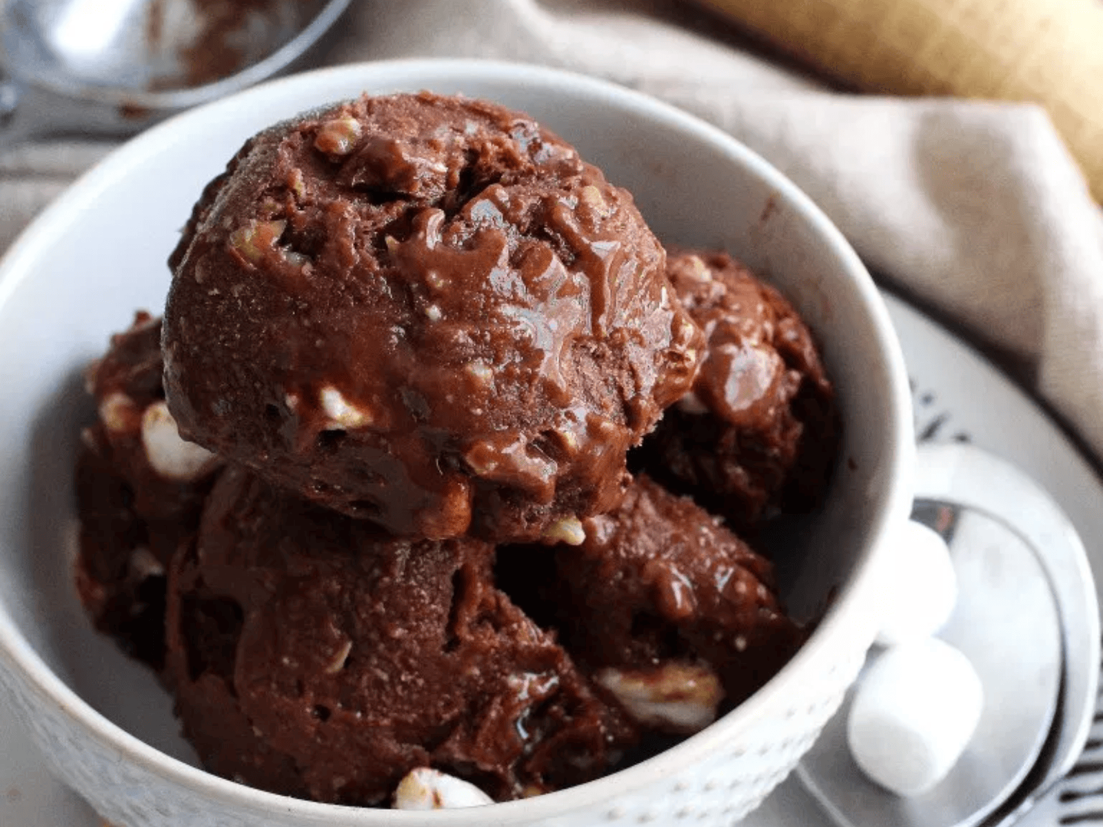 A scoop of creamy vegan Rocky Road ice cream, rich brown from cocoa, studded with white mini marshmallows and chopped roasted almonds, served in a bowl.