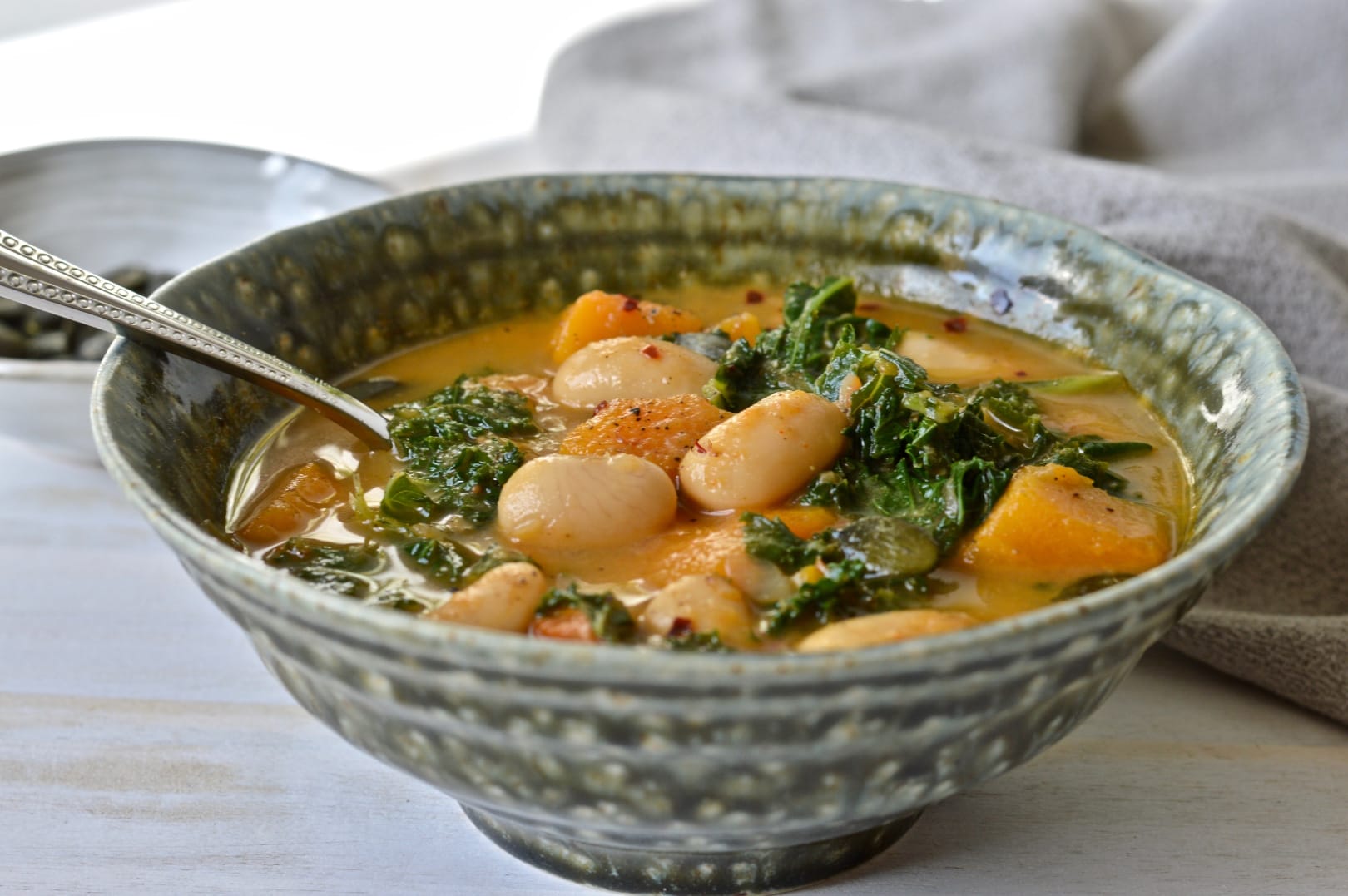 A rustic bowl of steaming vegan Bean, Kale and Butternut Stew, garnished with fresh herbs and served with a slice of crusty bread.