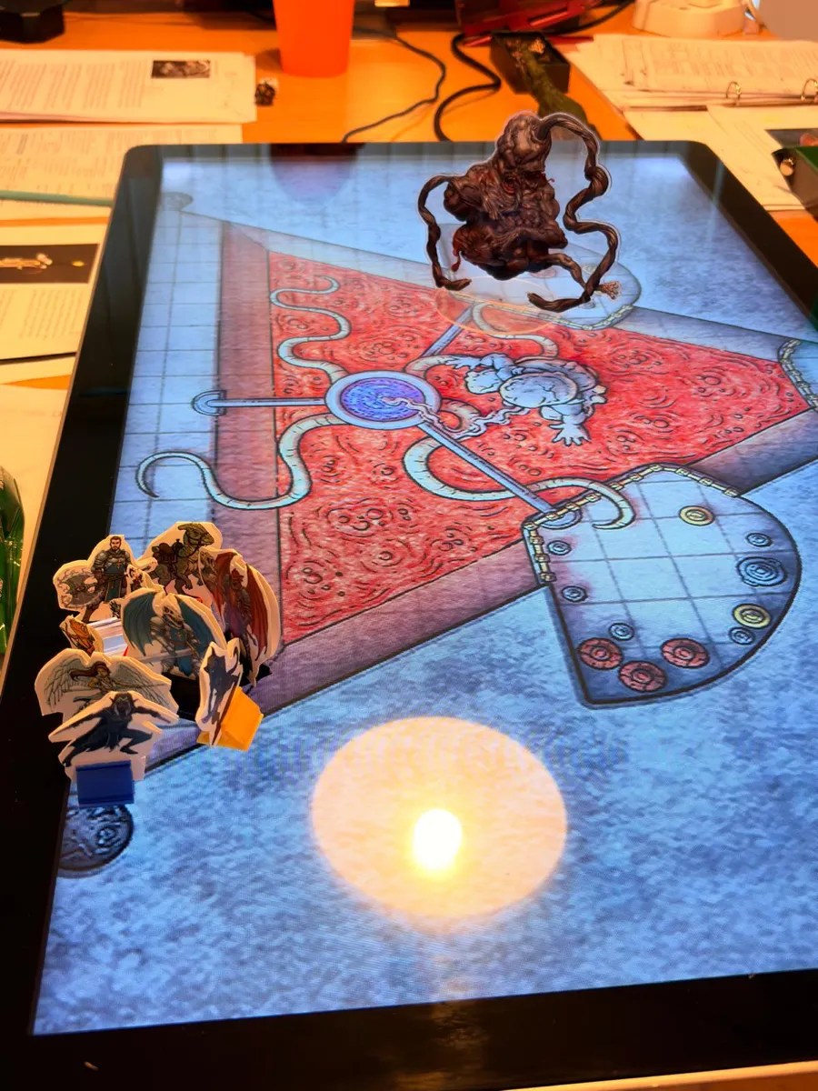 The atropal miniature hanging over the Area 77 map, with the team's standees grouped at the edge and a candle reflected in the screen.