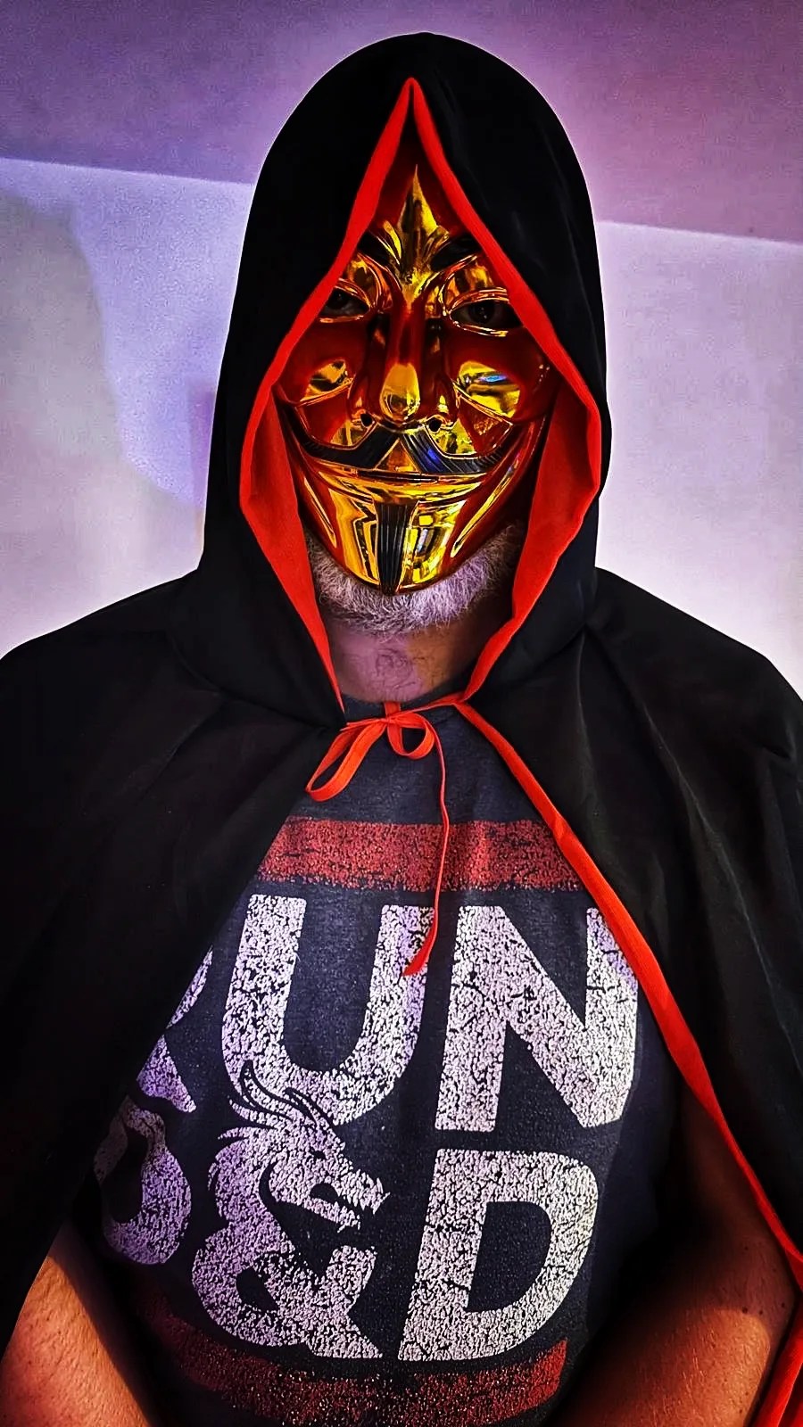 The DM in a black hooded cape and gold mask, wearing a Run D&D T-shirt.
