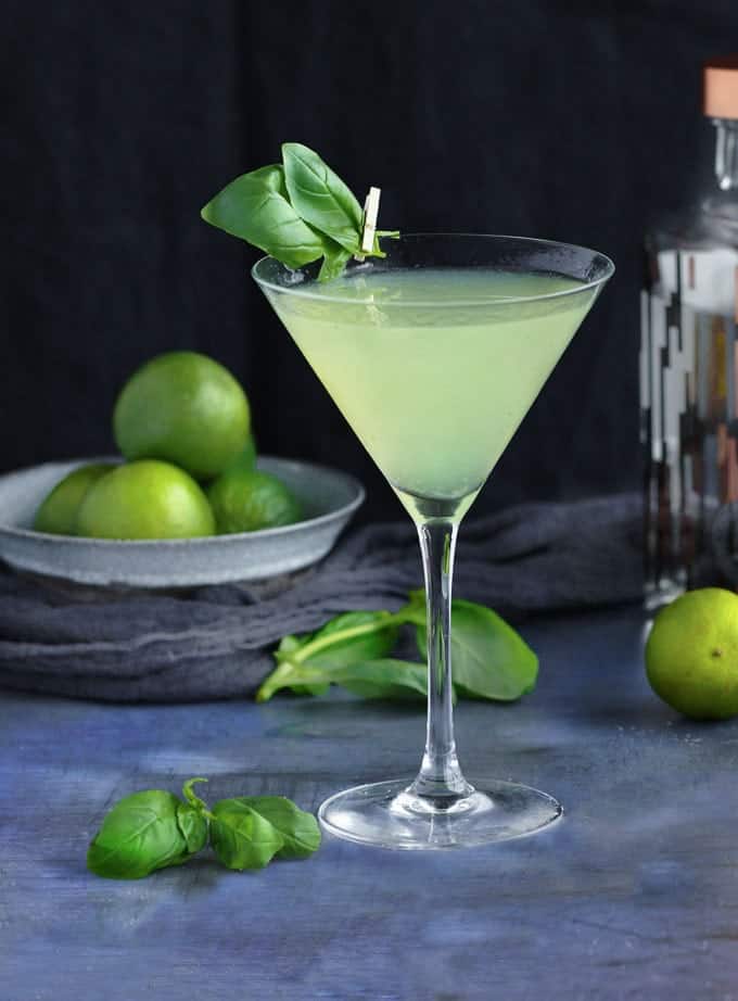 green hued cocktail in a martini glass with a basil leaf attached to the glass with a tiny clothespin and limes in the background