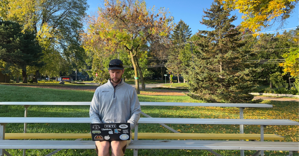 A picture of a man working remote on a bench in front of a baseball field