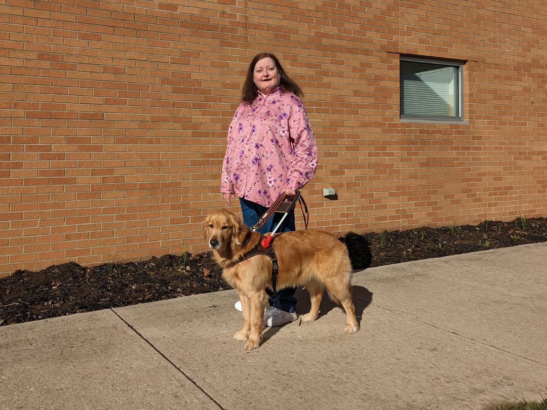 A smiling woman in a pink floral shirt stands in front of a brick wall with her new guide dog, a golden retriever. The dog is on her left and in harness.