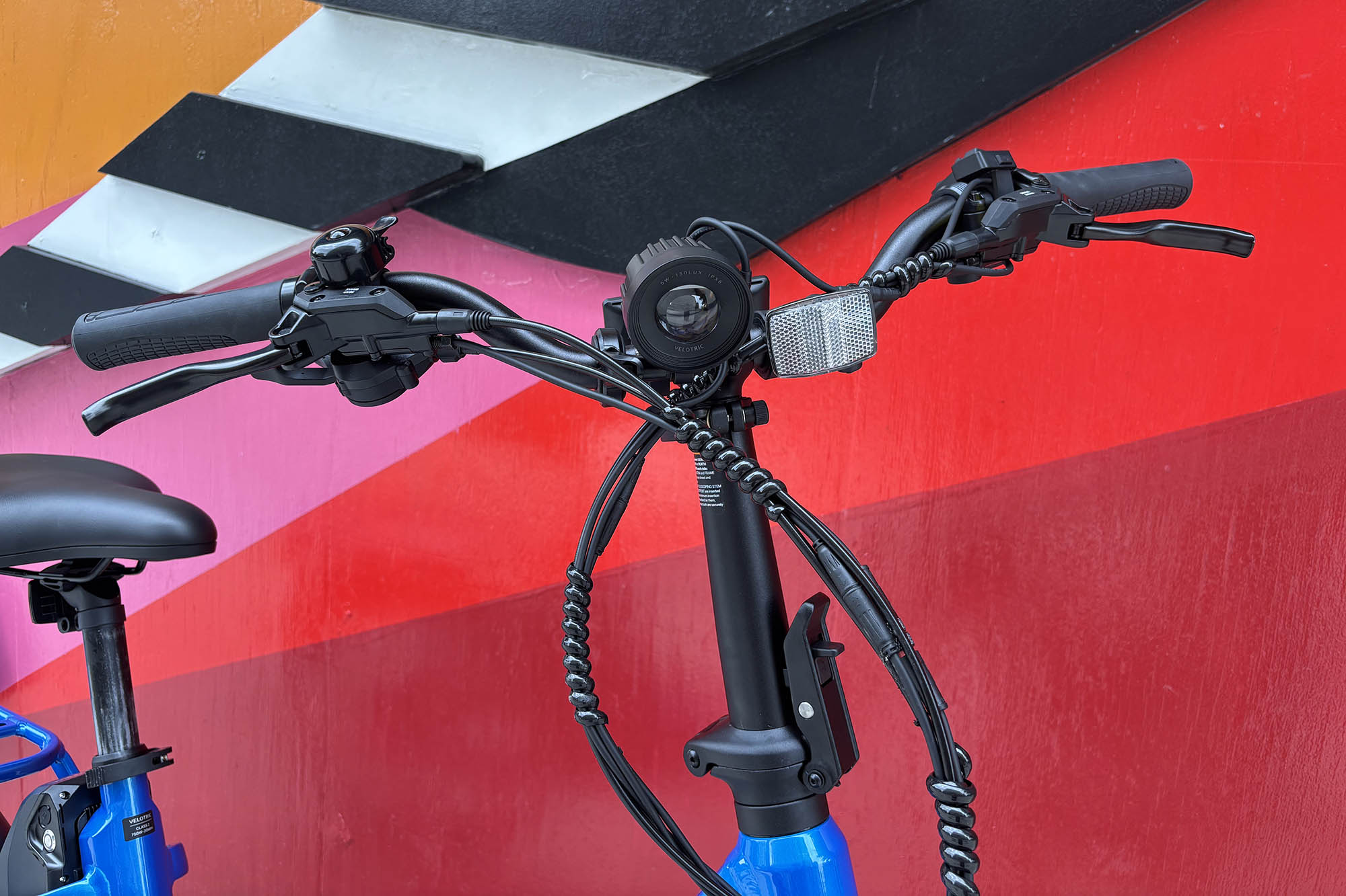 Closeup of the handlebars of a blue Velotric Fold 1 Plus folding ebike on its kickstand in front of a colorful mural