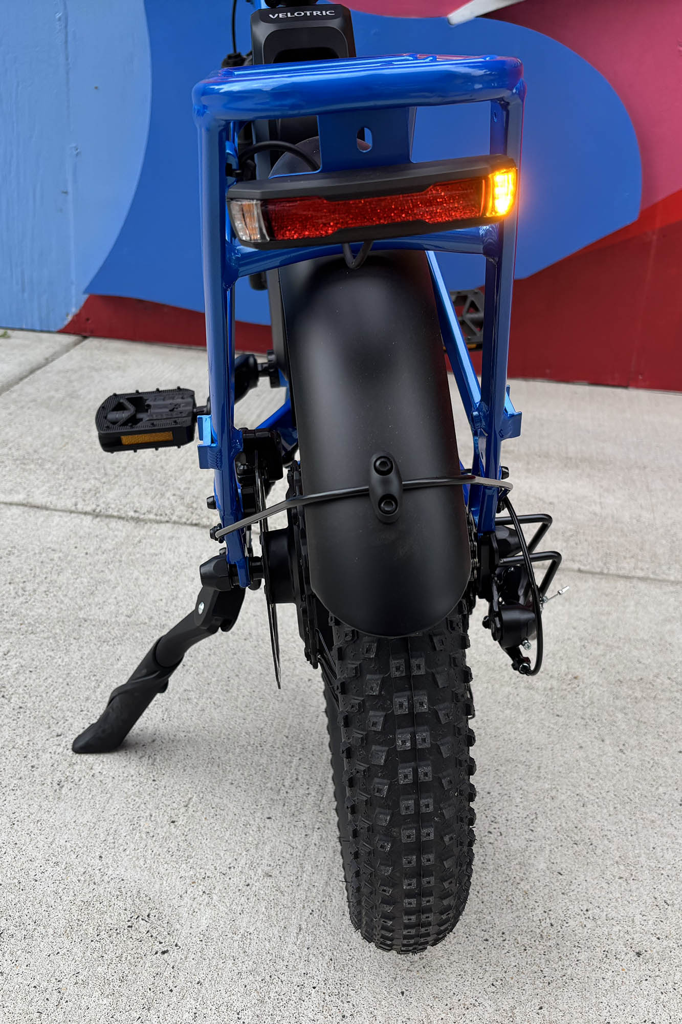 Closeup of the rear fender of a blue Velotric Fold 1 Plus folding ebike on its kickstand in front of a colorful mural