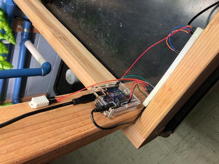 Arduino circuit board with control wires going from IO pins, and connected to USB and wall power, sitting on wooded shelving system for hydroponic garden, plastic piping for water in background