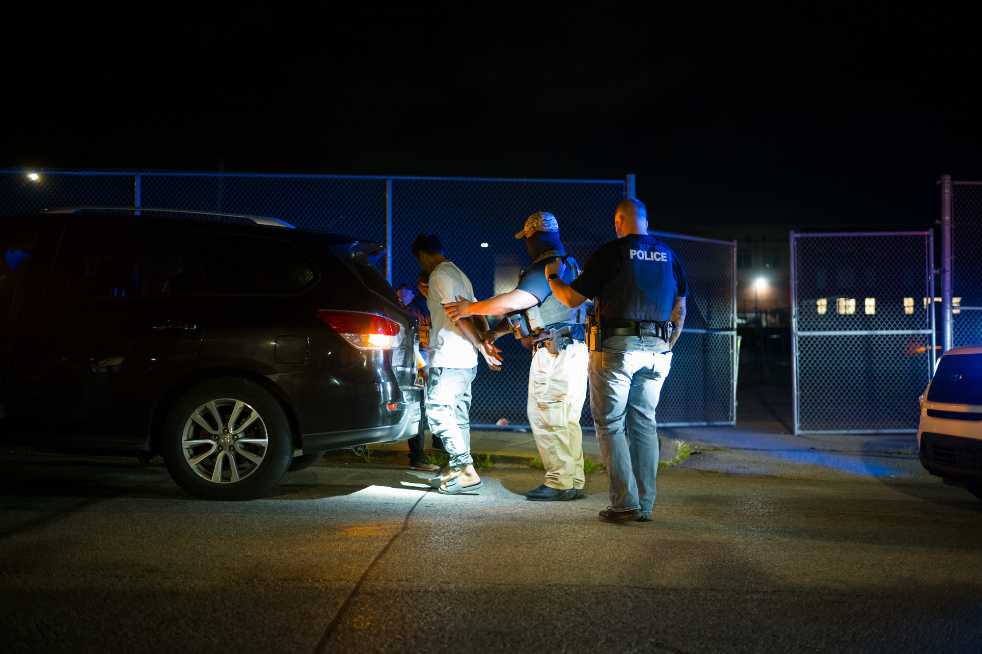 Two police officers detain and search a man next to a black SUV at night near a chain-link fence, illuminated by artificial light.