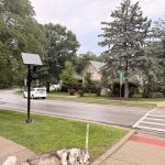 A Flock camera pole with solar energy panels above it stands on the parkway next to a street. A house and some trees are visible behind it.