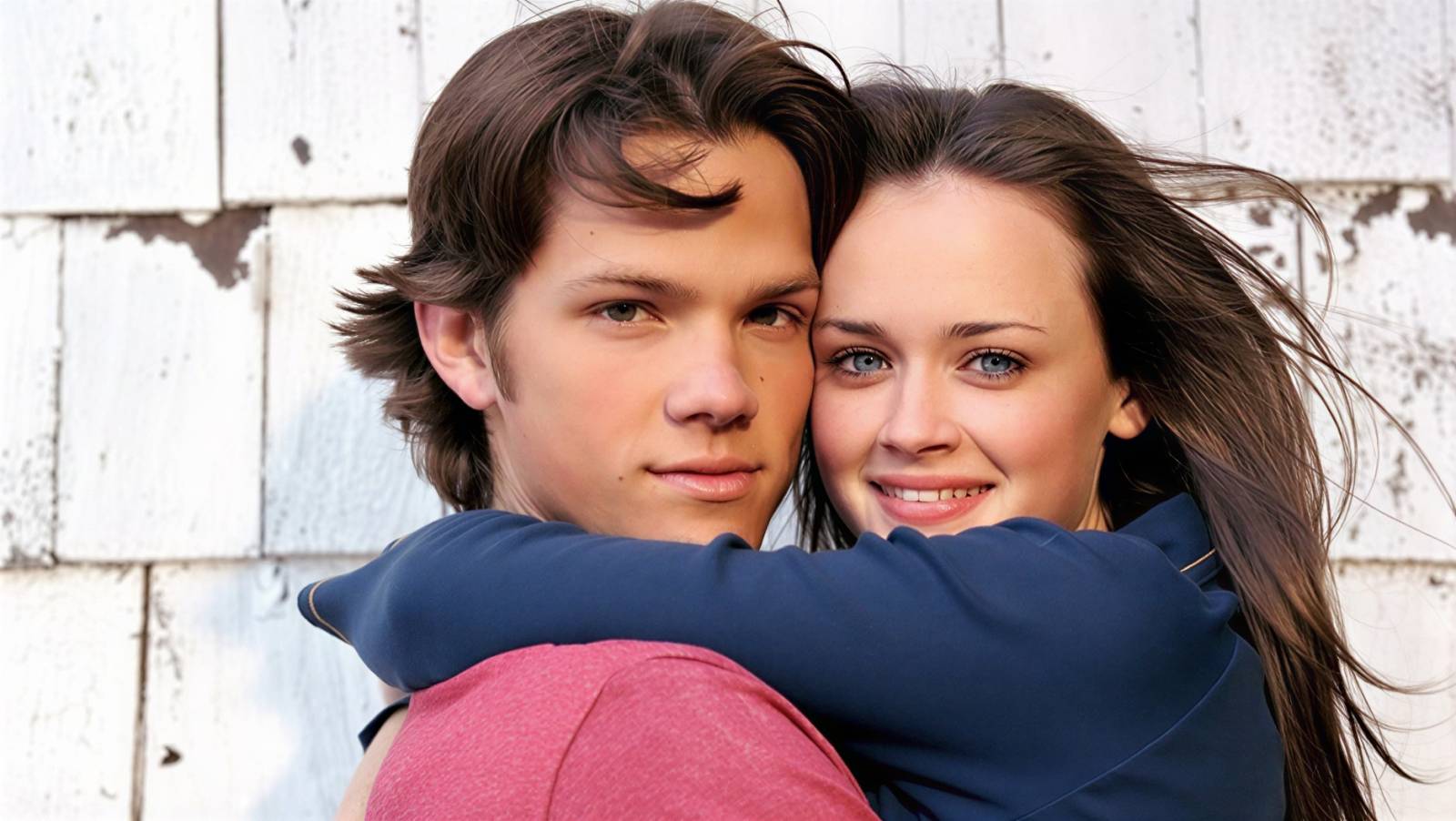 Young couple hugging and smiling at the camera in front of a weathered white wooden wall.